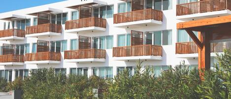 Un hotel moderno con varios balcones, ventanas amplias y abundante vegetación en primer plano.