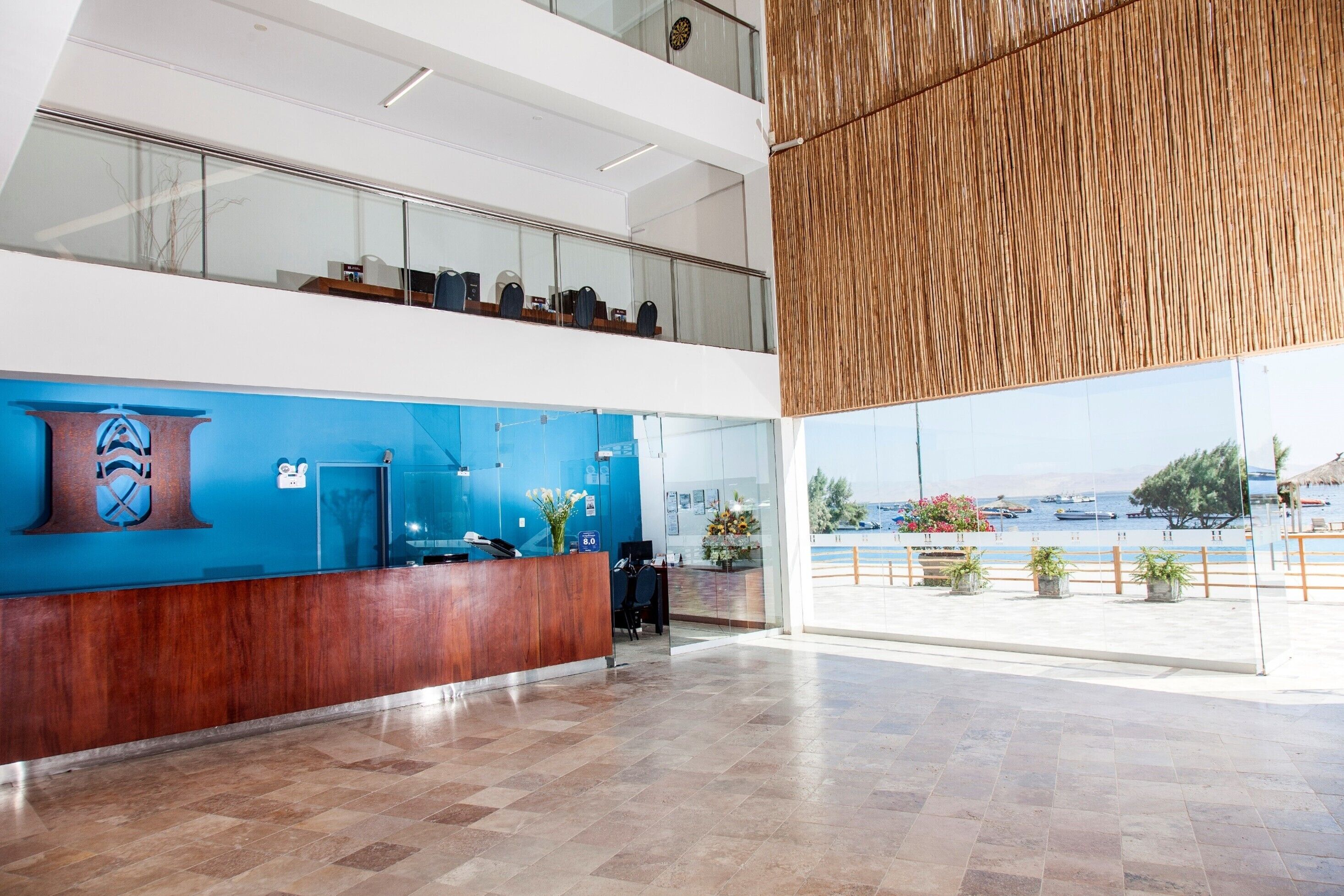 Un área de recepción moderna con un mostrador de madera, una pared azul con un logo y vistas al mar a través de amplios ventanales.