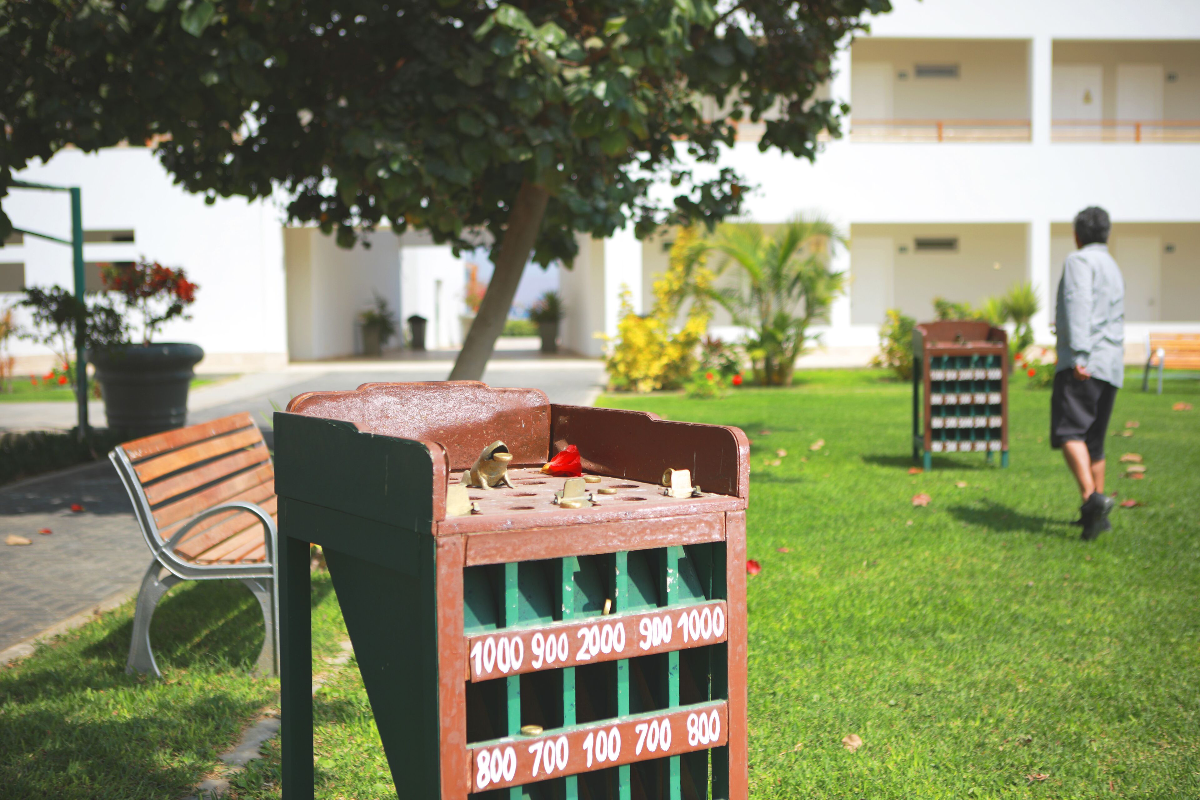 Un cubo de basura verde con ranuras numeradas, un banco y un edificio al fondo.
