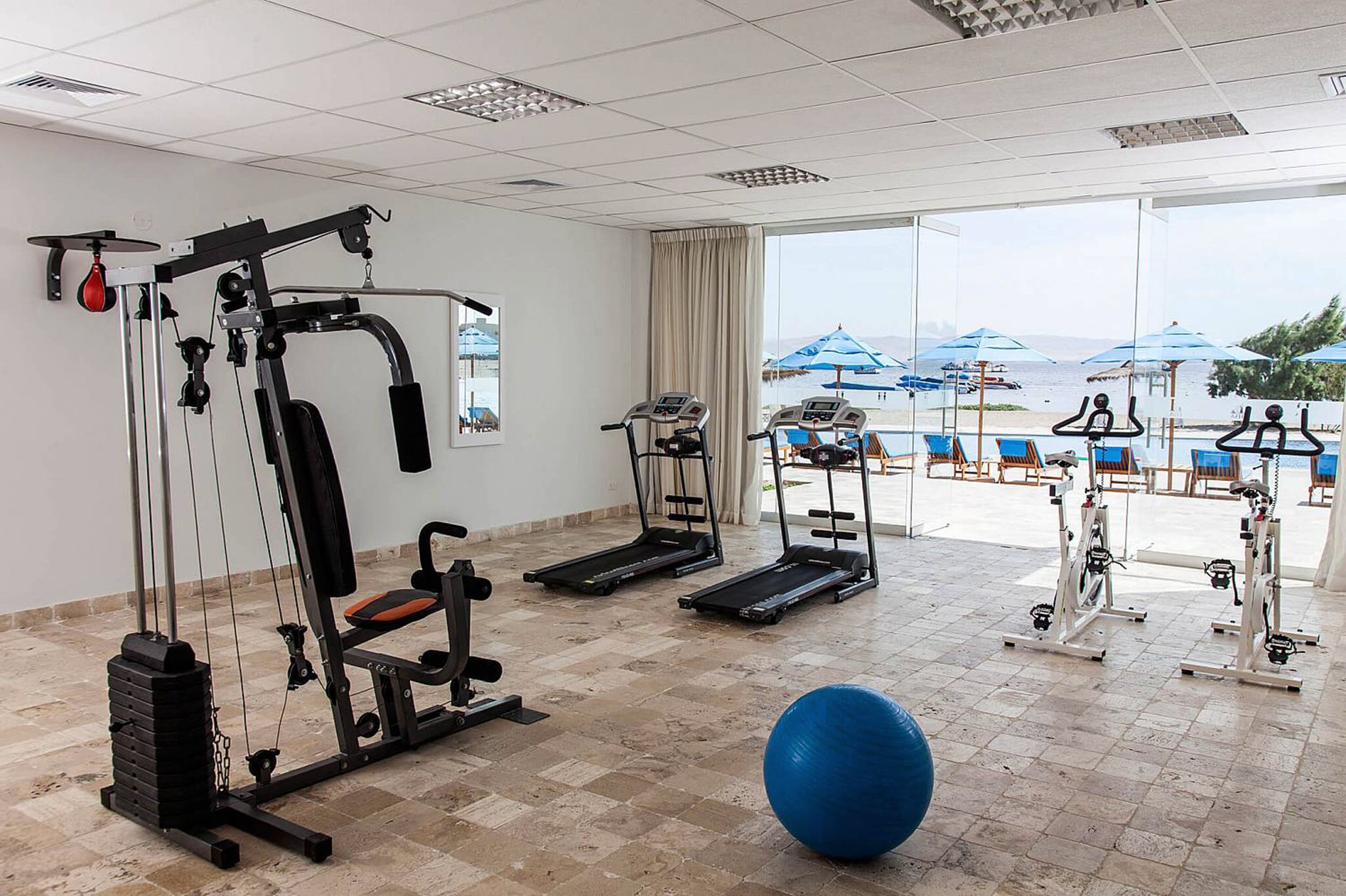 Un gimnasio con equipo de ejercicio, una pelota de ejercicio azul y vista a una playa con sombrillas.