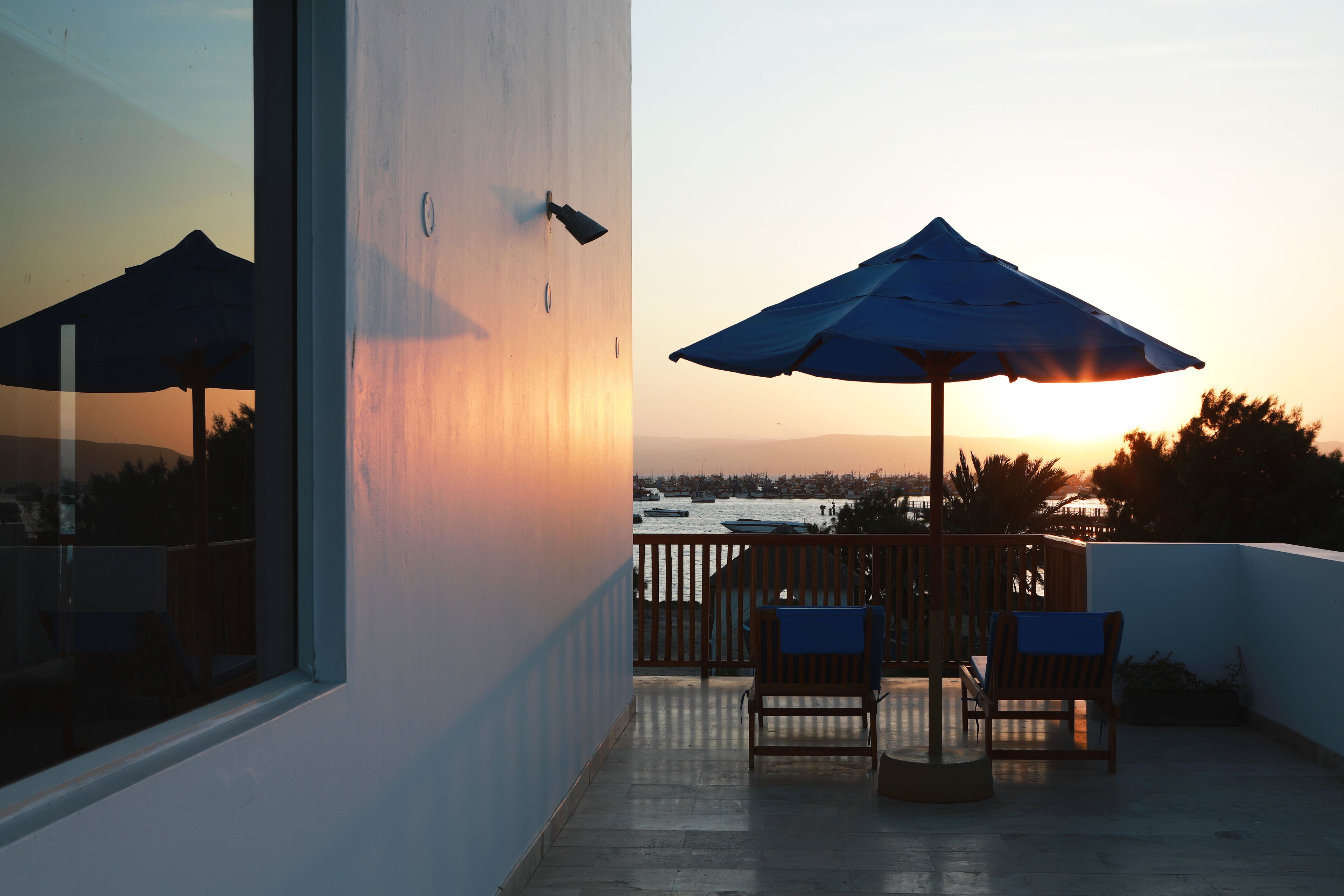 A balcony with a table and chairs, a large umbrella, and a view of the sunset over the water.