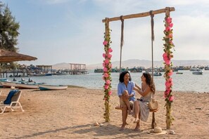 A beach setting with a swing decorated with pink flowers, a wooden structure, and boats in the background. - Hotel San Agustín Paracas (Paracas)