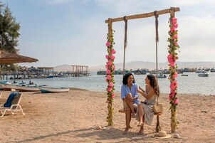 Un paisaje de playa con una hamaca adornada con flores rosadas, una estructura de madera y embarcaciones al fondo.