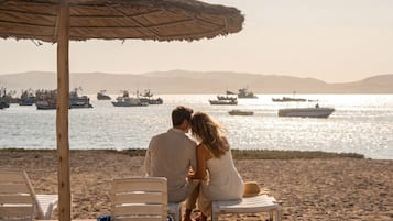 Una coppia seduta su una sedia da spiaggia sotto un ombrellone di paglia, con vista su un mare calmo e imbarcazioni.
