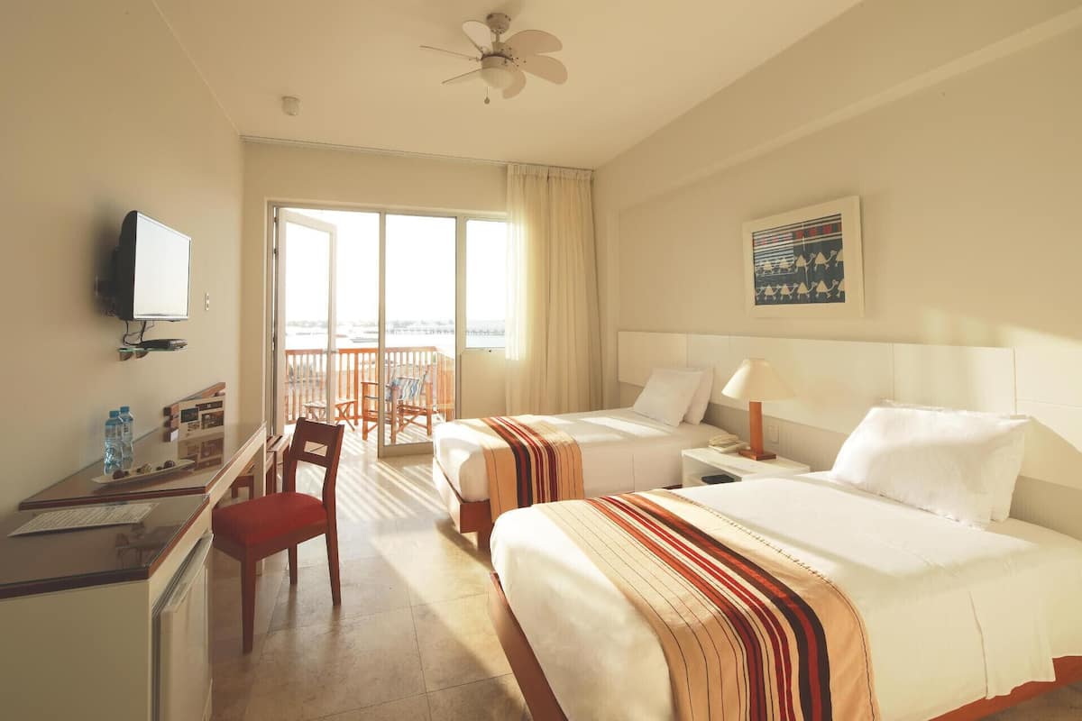 A hotel room with two beds, a desk with a chair, a television, and a balcony with a view.
