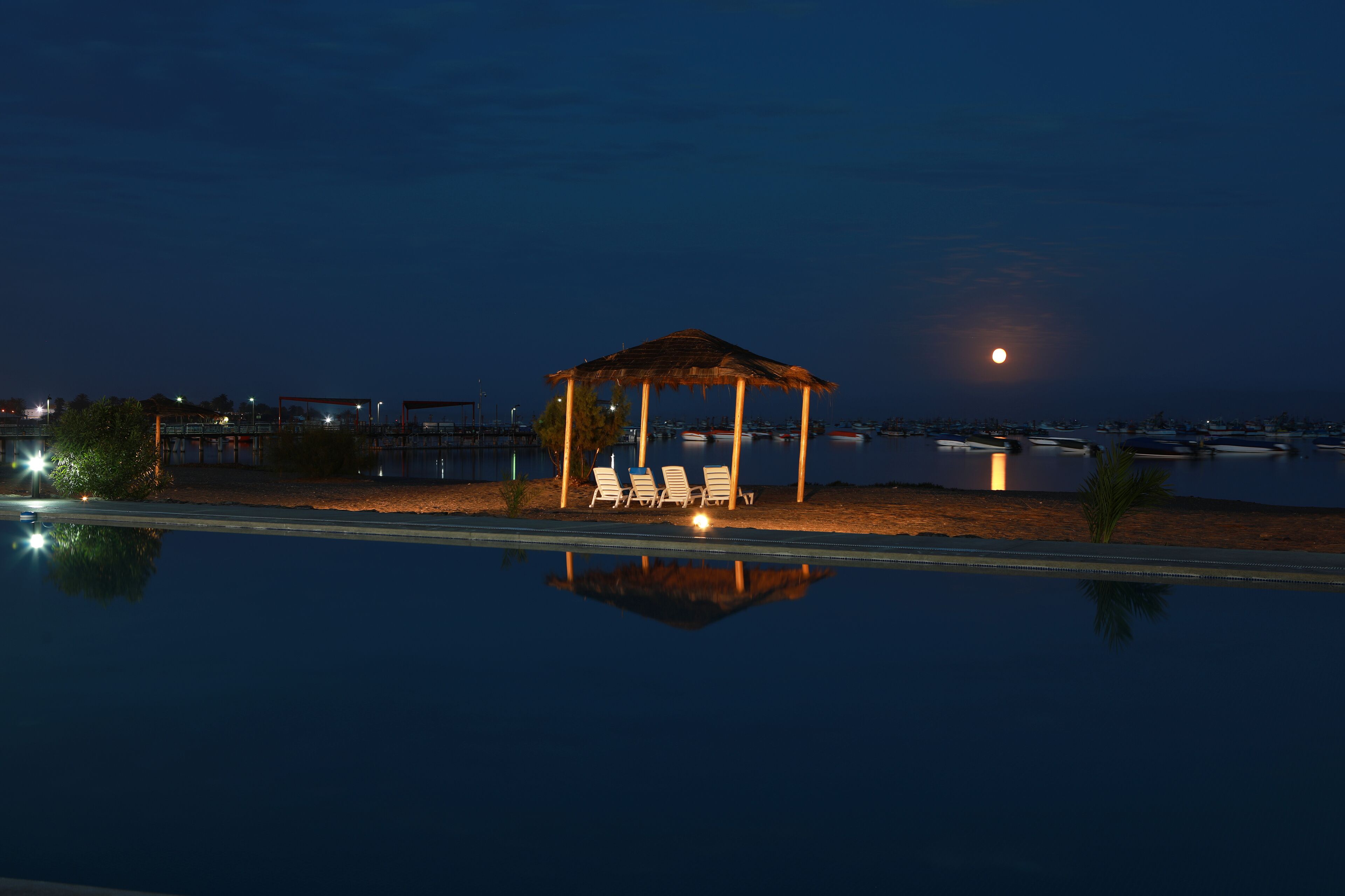 A nighttime pool area with a gazebo and lounge chairs, reflecting the moon and surrounding buildings.
