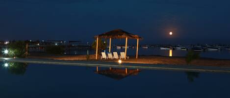 Zona de piscina nocturna con un pabellón y sillones de descanso, reflejando la luna y edificios circundantes.