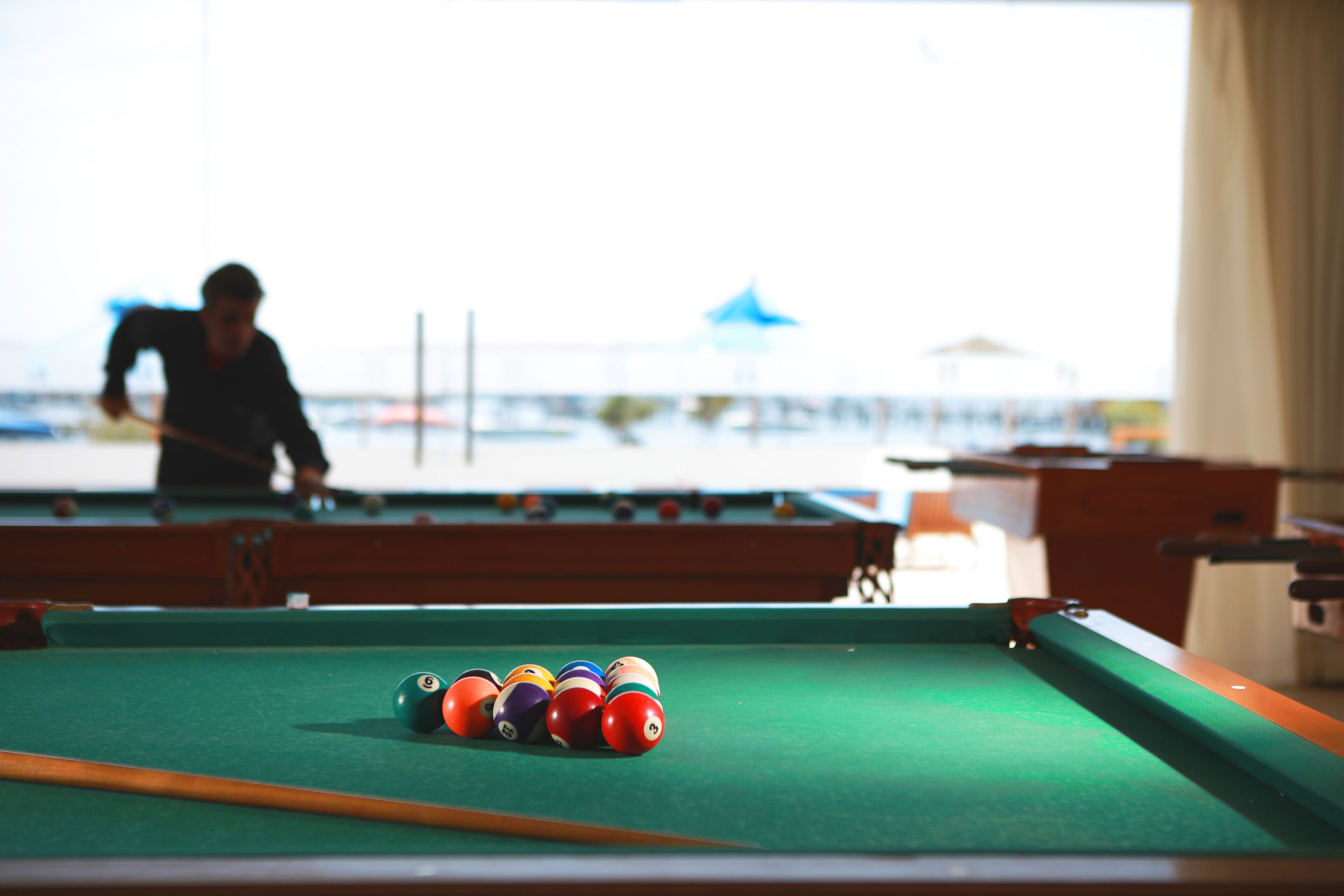 A pool table with balls and a cue stick, a person in the background, and a view of the sea.