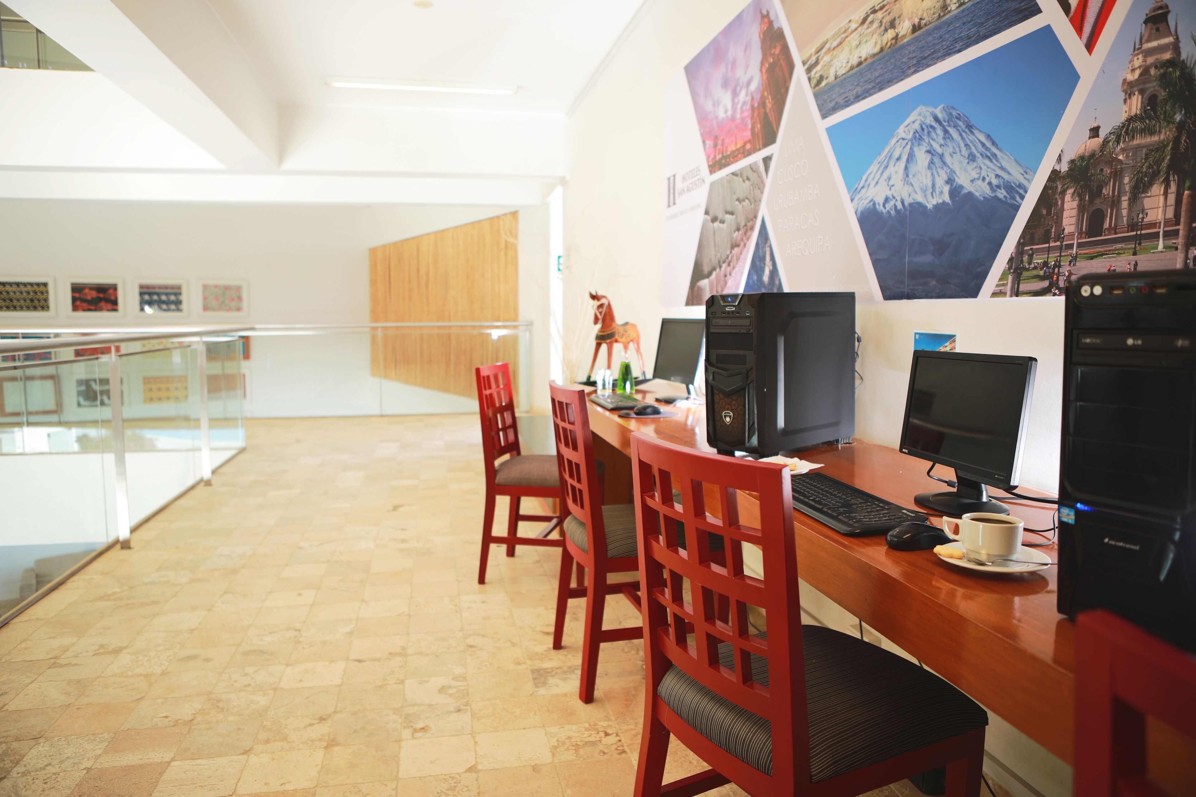 A modern office space with a wooden desk, two computer monitors, a keyboard, and a cup of coffee. There are red chairs and a glass railing in the background.