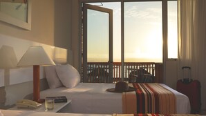 A hotel room with a bed, a nightstand, a lamp, a suitcase, and a view of the sunset through a large window. - Hotel San Agustín Paracas (Paracas)