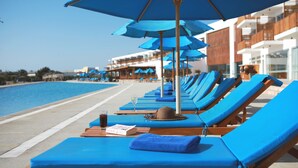 A row of blue lounge chairs with umbrellas by a poolside, wooden tables with drinks and a hat, and a building with balconies in the background. - Hotel San Agustín Paracas (Paracas)