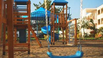 Explore the playground with slides and swings, set amidst a lush garden with palm trees and colorful flowers.
