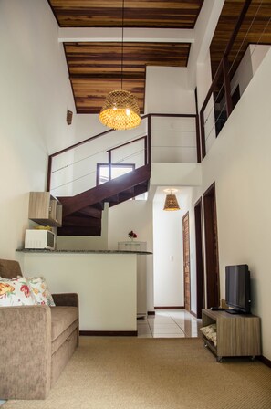A spacious living area with a high ceiling, a wooden staircase, a kitchen area, and a television on a wooden stand.