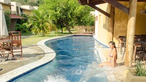 A swimming pool with a person swimming, surrounded by a covered patio area with chairs and tables. - Bosque da Praia - ePipa Hotéis (Tibau do Sul)
