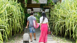 A couple walking towards a building labeled "Bosque da Praia" with a suitcase and a pink bag. - Bosque da Praia - ePipa Hotéis (Tibau do Sul)