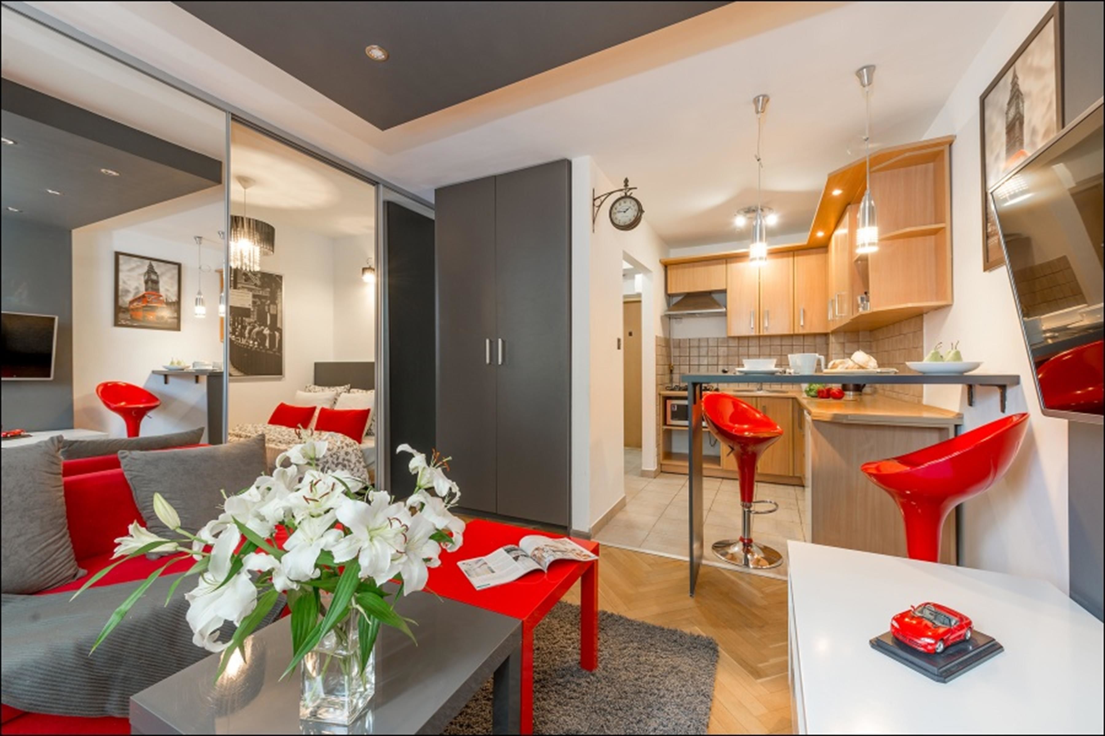 A modern living room with a red sofa, a dining table, and a kitchen area with wooden cabinets and red bar stools.