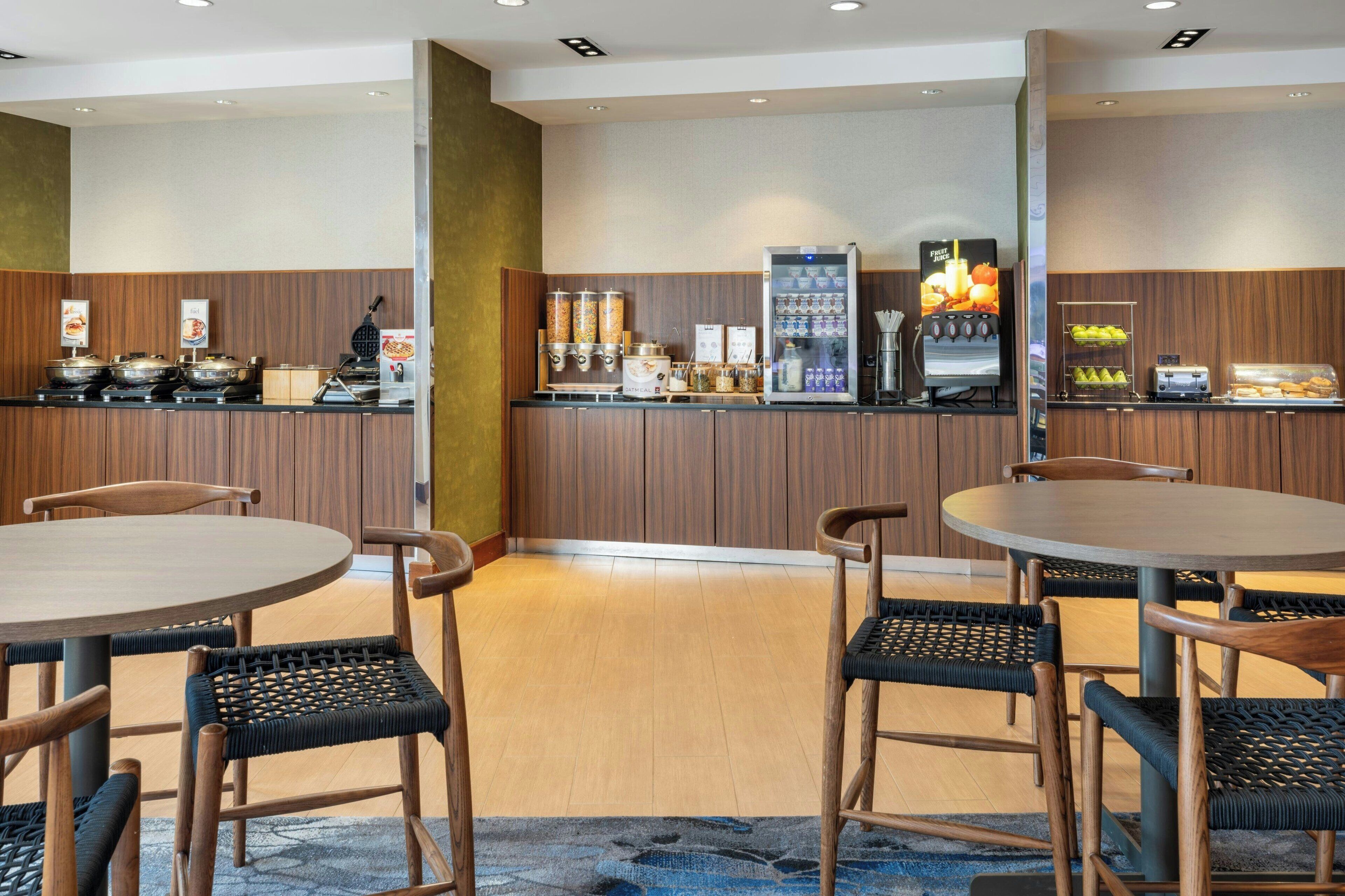 A hotel dining area with wooden panel walls, a buffet counter with various food items, a beverage dispenser, and round tables with wicker chairs.