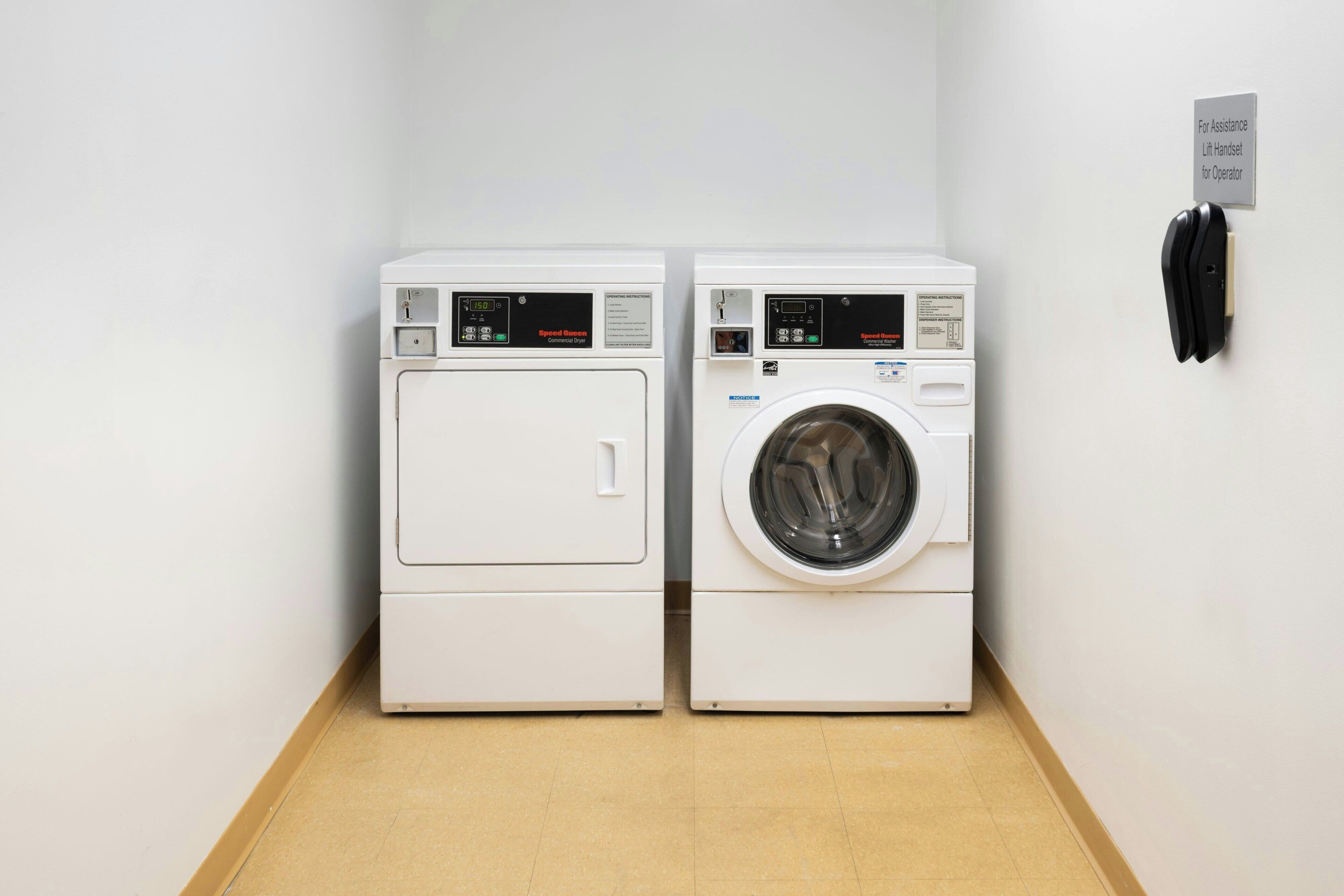A laundry area with two white washing machines and a dryer, a sign for assistance, and a handrail.