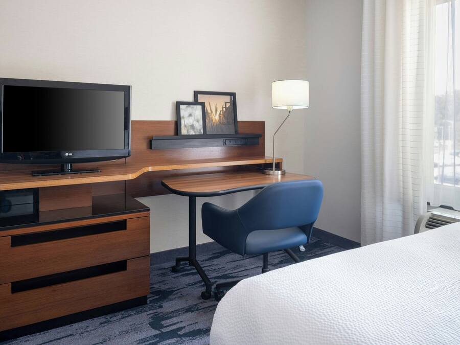 A hotel room with a bed, a desk with a flat-screen TV, a chair, a lamp, and a window with curtains.