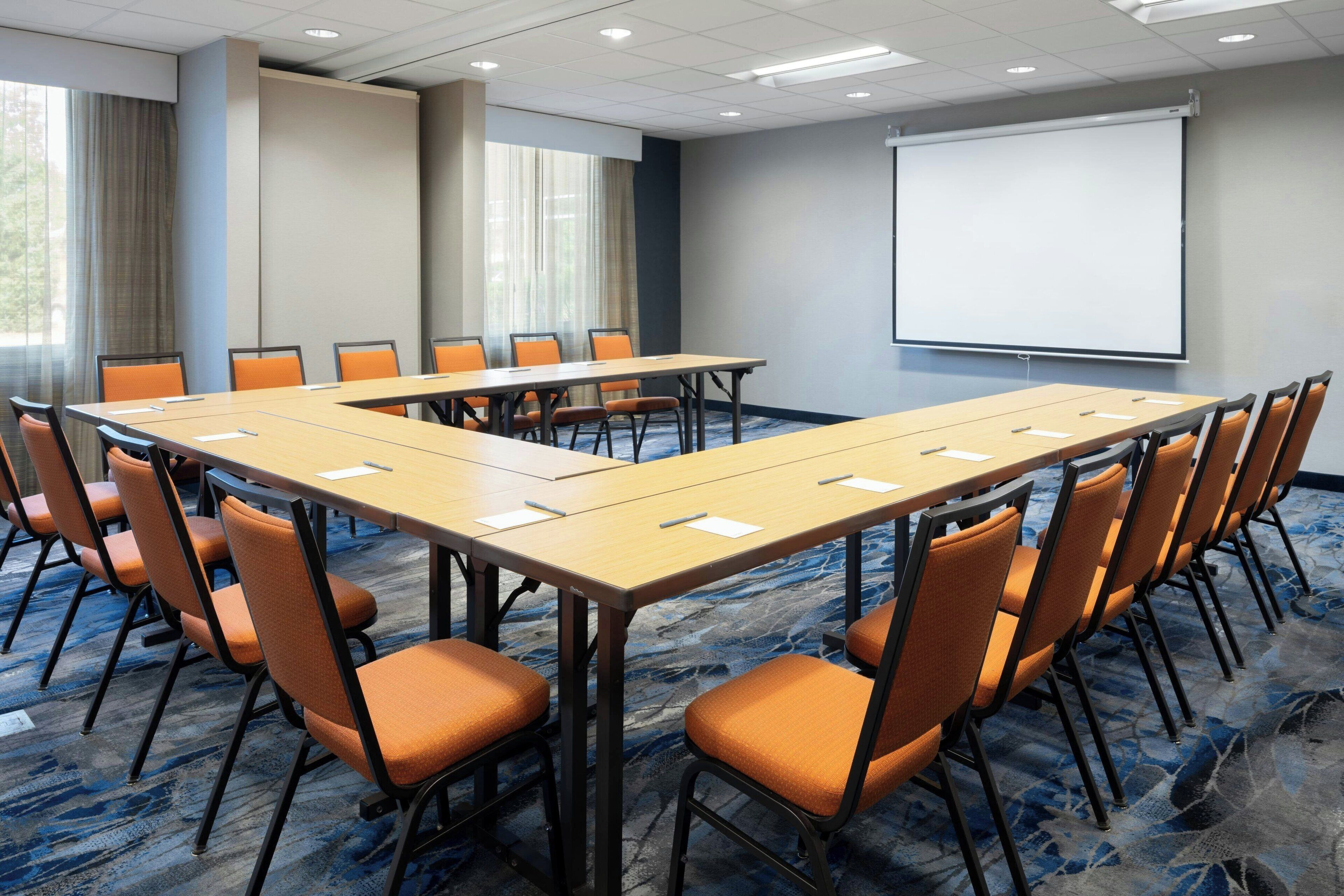 A conference room with a long table, orange chairs, a projector screen, and a carpeted floor.