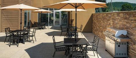 An outdoor patio with tables and chairs, umbrellas, and a barbecue grill.