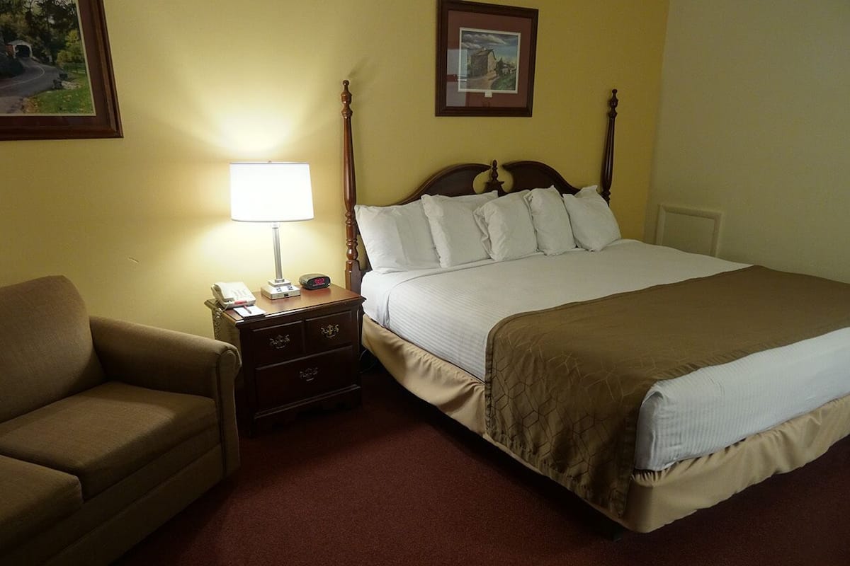 A hotel room with a bed, a bedside table, a lamp, a chair, and a picture on the wall.