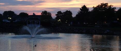 A serene evening by a lake with a fountain, surrounded by trees and a gazebo.