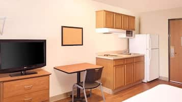 A compact hotel room with a bed, a TV, a small dining table, a chair, a kitchenette with a microwave and refrigerator, and wooden cabinets.