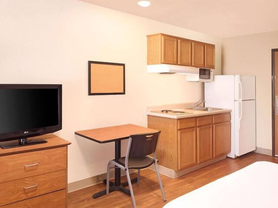 A compact hotel room with a bed, a TV, a small dining table, a chair, a kitchenette with a microwave and refrigerator, and wooden cabinets.