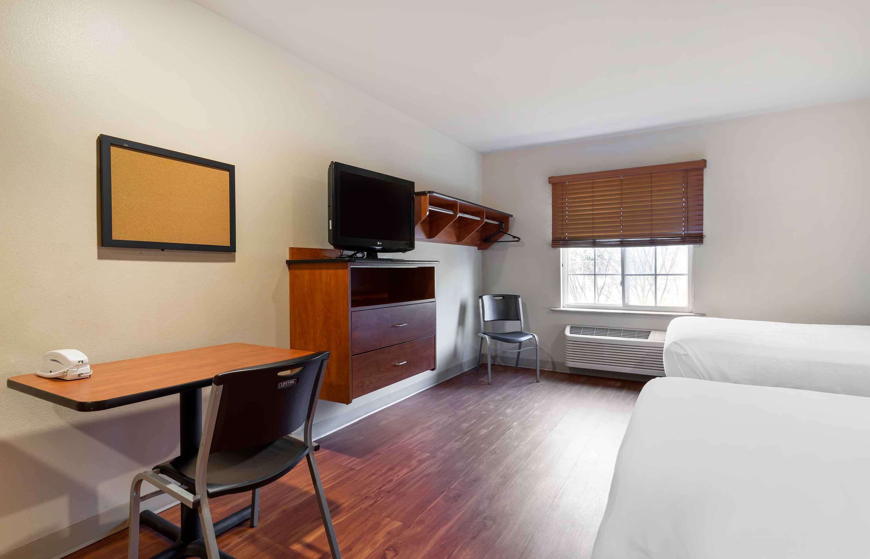 A hotel room with a bed, a desk with a chair, a television, a small table, a chair, a window with blinds, and a picture frame on the wall.