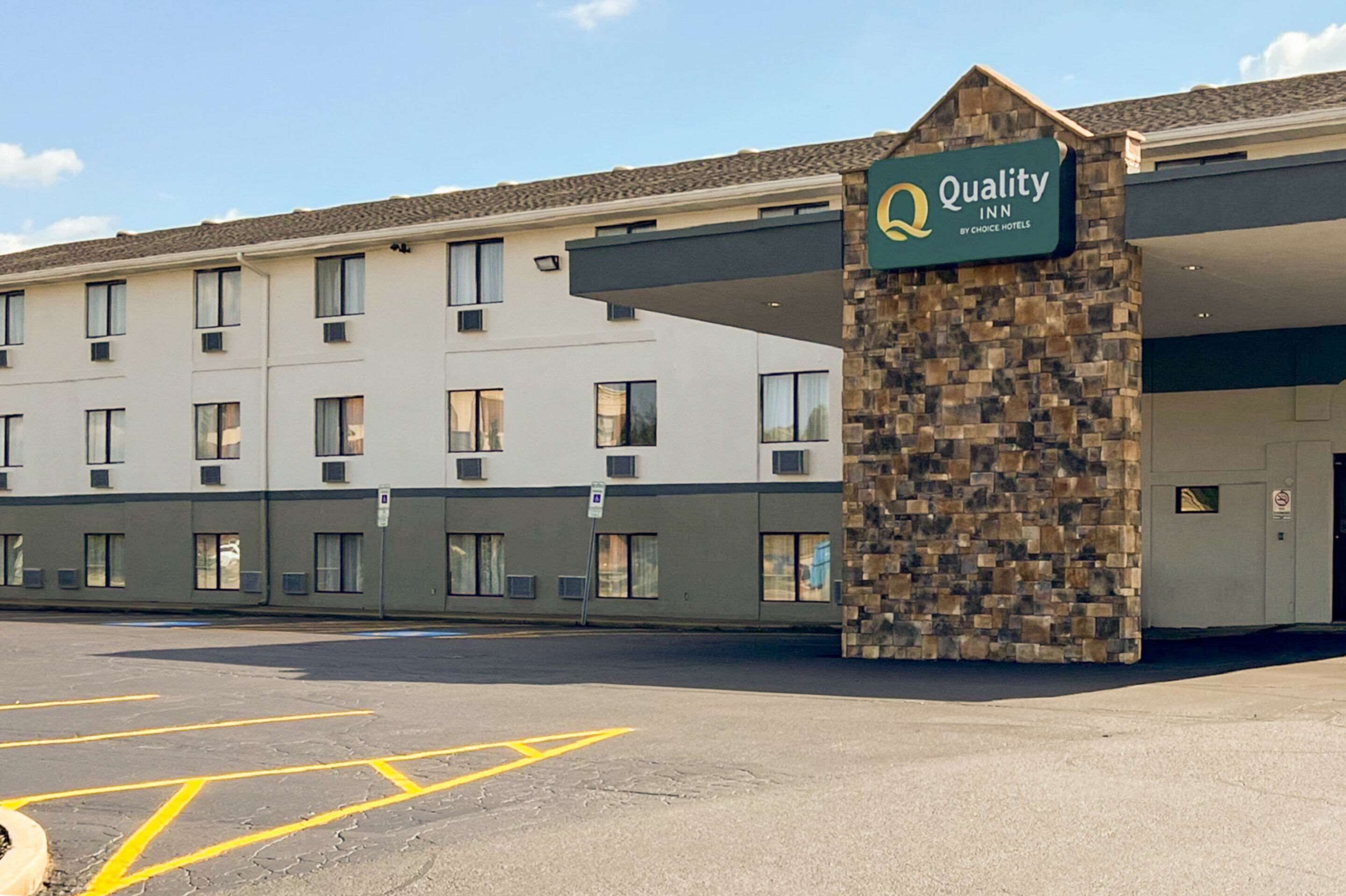 L’entrée d’un hôtel, avec une enseigne indiquant « Quality Inn by Choice Hotels », un parking et la façade d’un immeuble dotée de nombreuses fenêtres et d’un pilier en pierre.