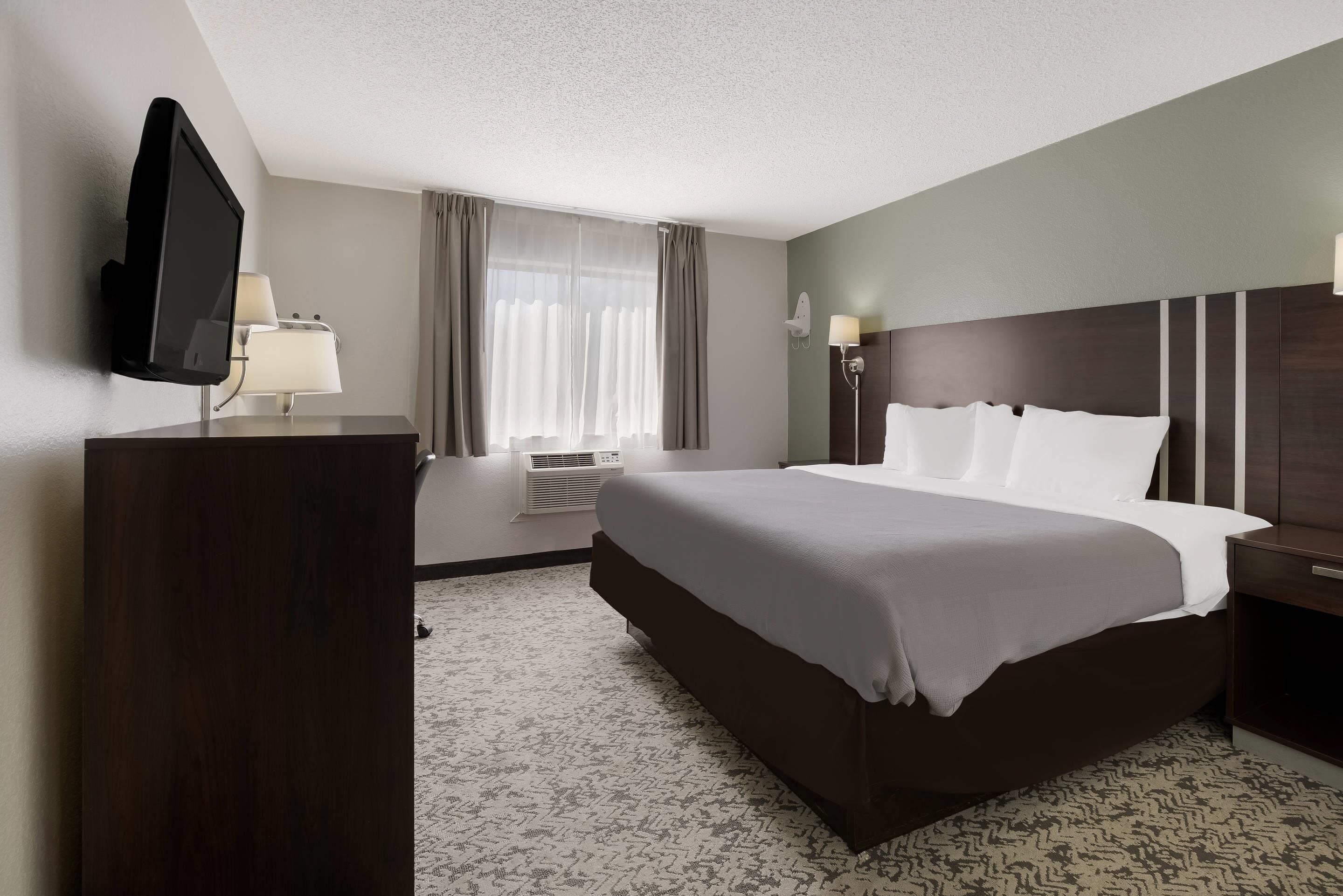 A hotel room with a large bed, a flat-screen TV mounted on the wall, a desk with a lamp, and a window with curtains.