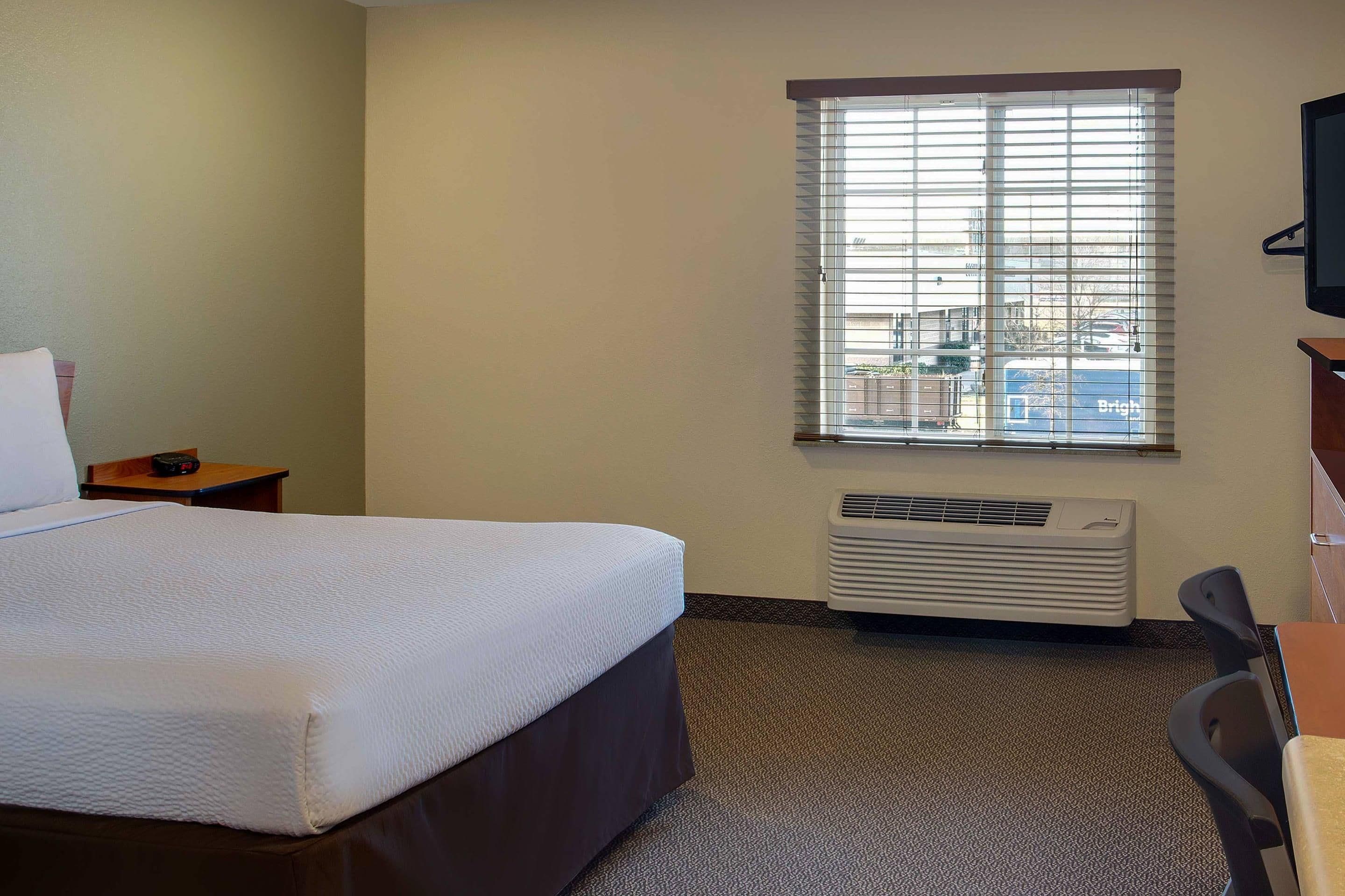Habitación de hotel con cama, escritorio, silla, ventana con persianas y unidad de aire acondicionado.