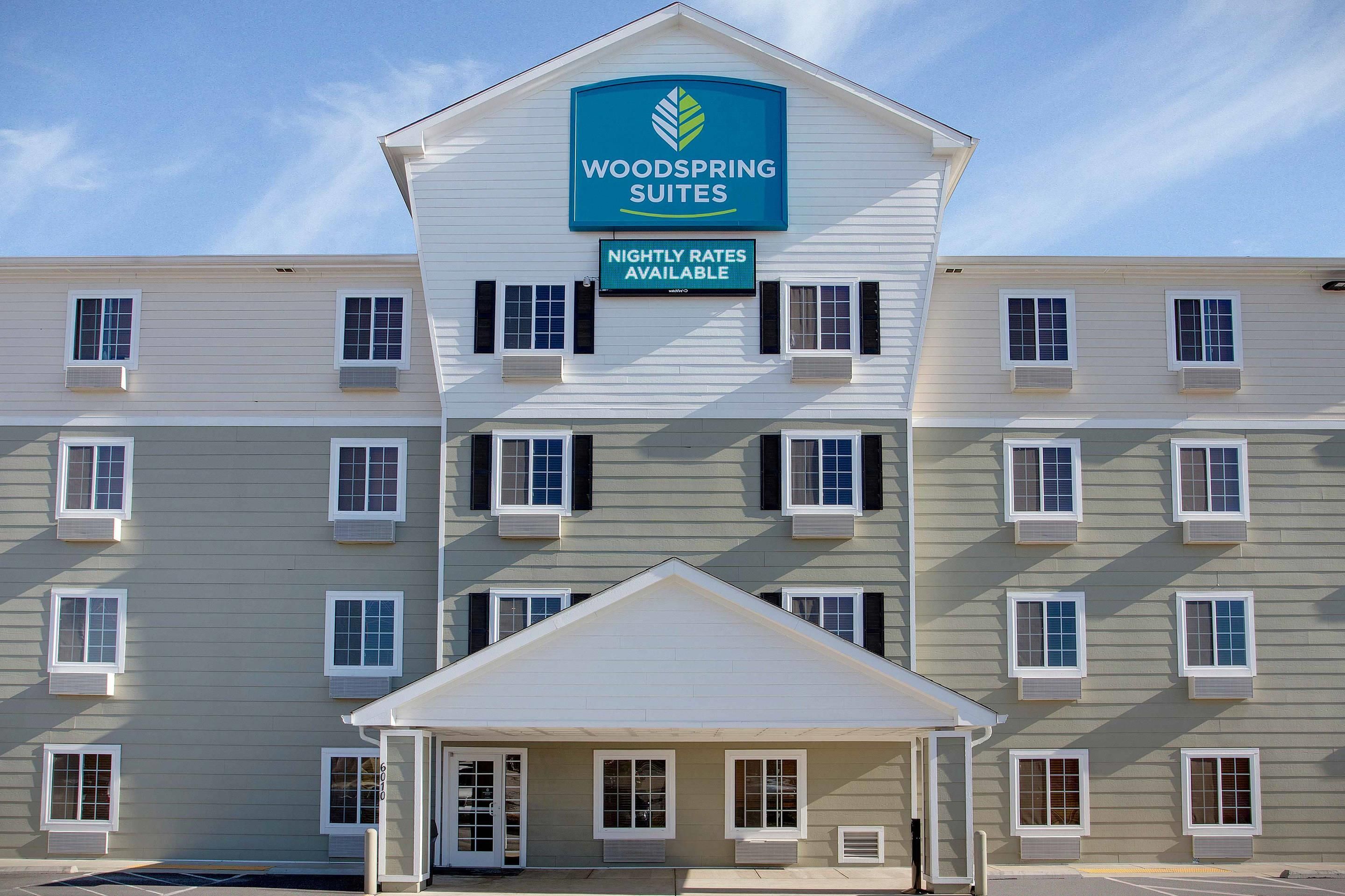 Um hotel com vários andares e uma placa de destaque indicando 'Woodspring Suites' e 'Tarifas por Noite Disponíveis'.