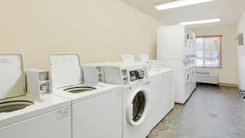 Une buanderie équipée de plusieurs machines à laver et de plusieurs sèche-linge, dotée d’un sol carrelé et d’une fenêtre avec des stores.