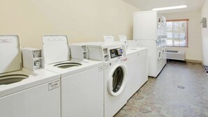 A laundry room with multiple washing machines and dryers, a tiled floor, and a window with blinds. - WoodSpring Suites Washington DC Andrews AFB (Suitland)