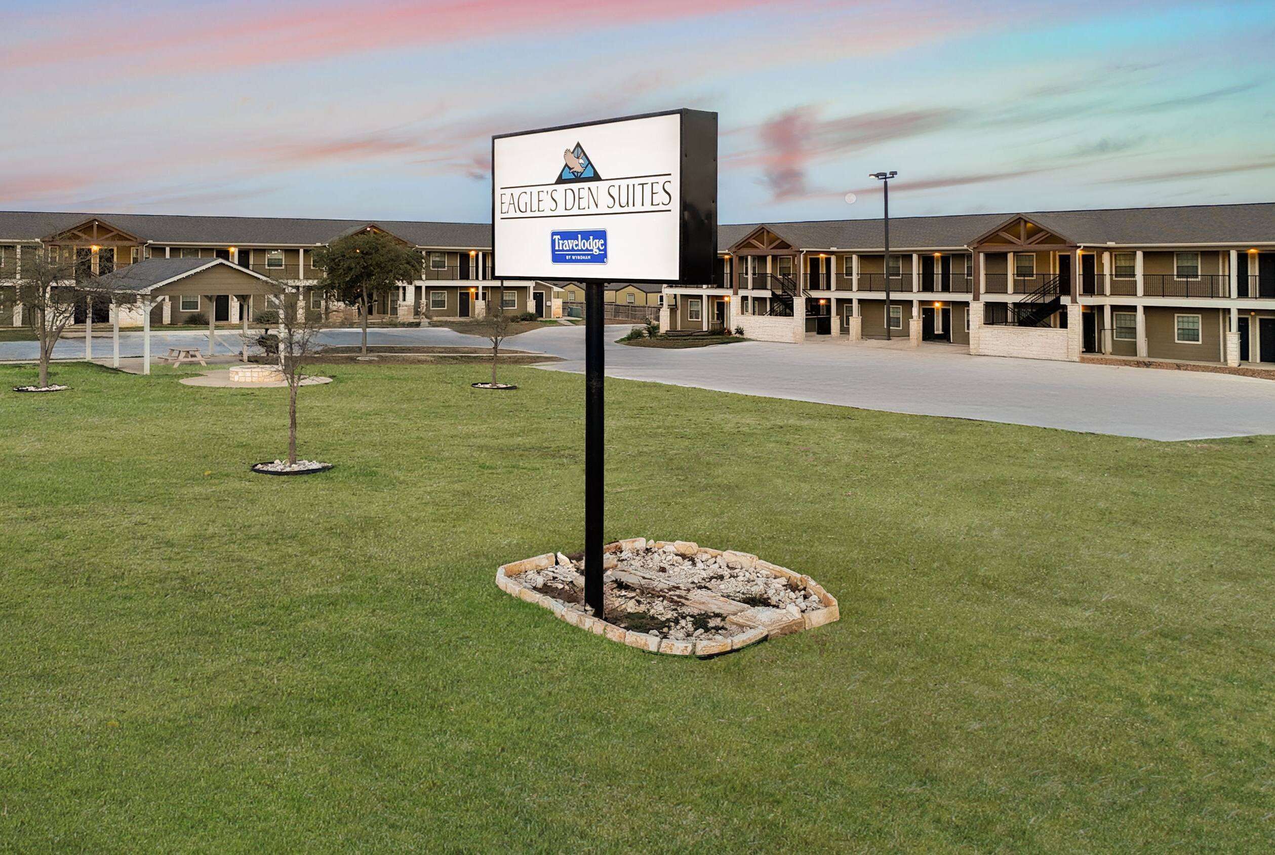 A Travelodge sign in front of Eagle's Den Suites, a multi-story motel with a central courtyard and outdoor seating area.