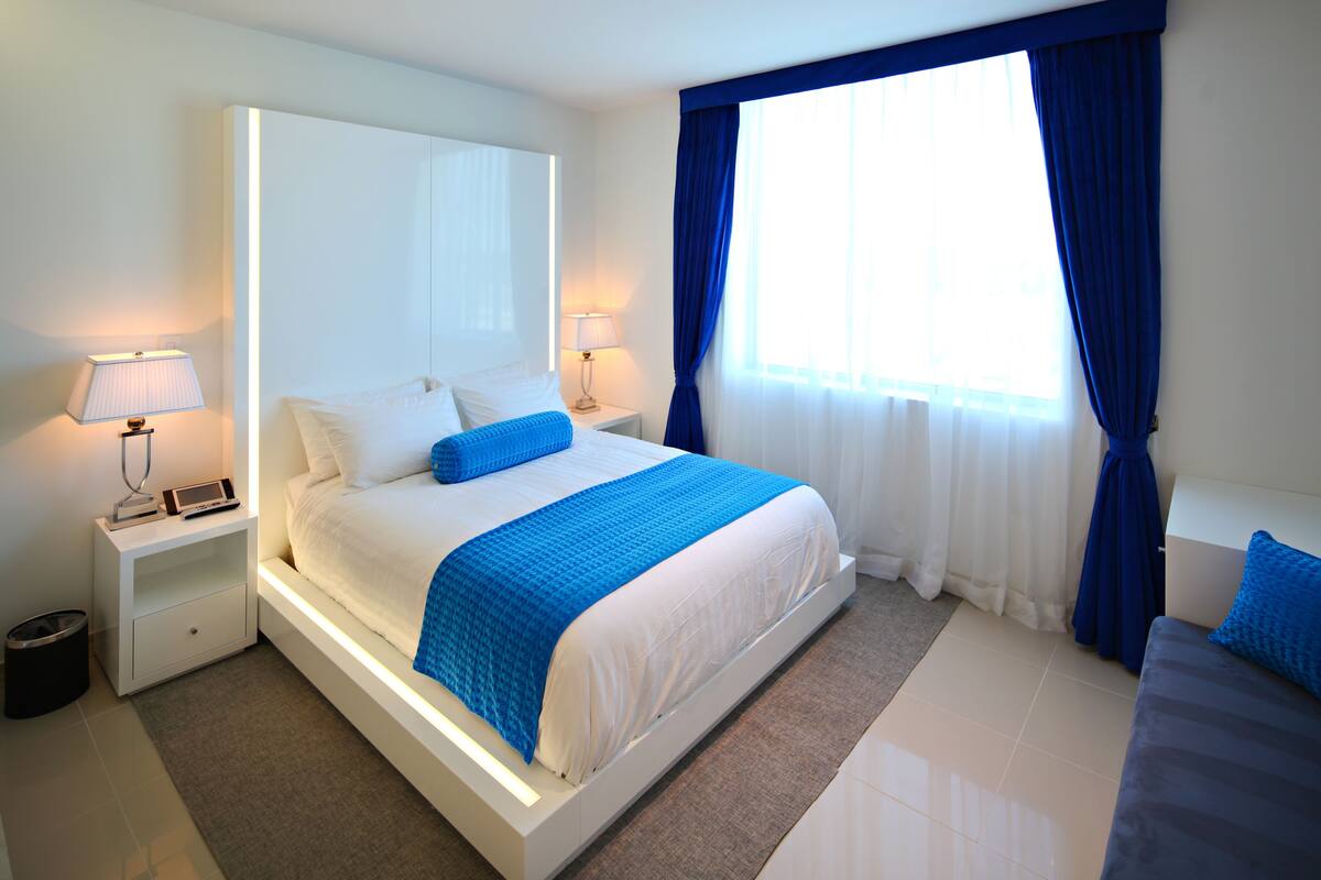A modern hotel room with a large bed, bedside lamps, a nightstand, and a view of the outdoors through a window with blue curtains.