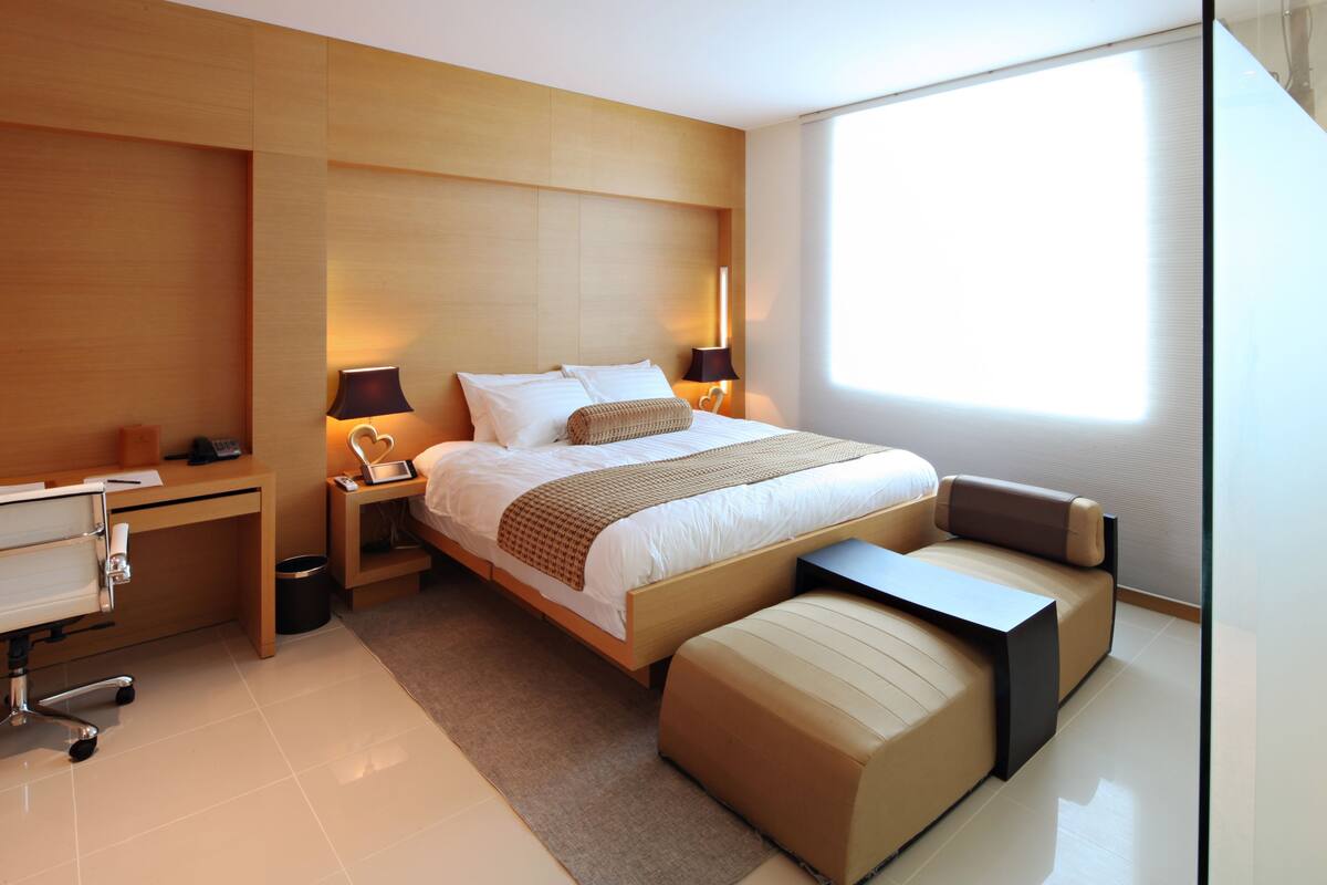 A modern hotel room with a large bed, bedside lamps, a desk, and a window.