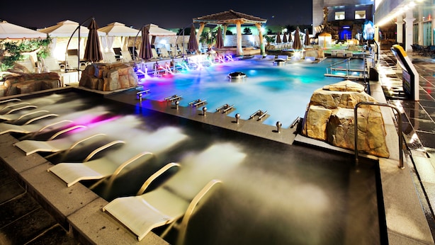Uma área de piscina à noite, com espreguiçadeiras, hidromassagem e um gazebo.