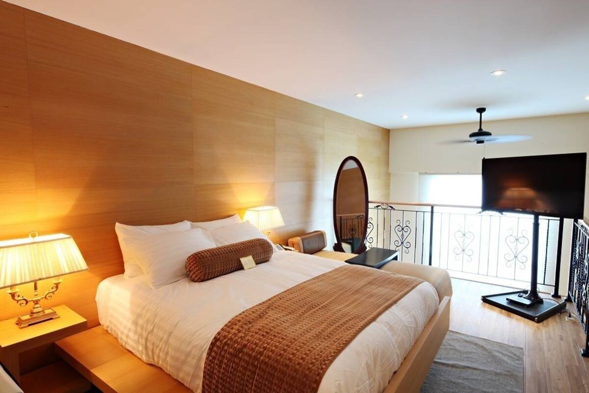 A hotel room with a large bed, two bedside lamps, a flat-screen TV, and a balcony with a railing.
