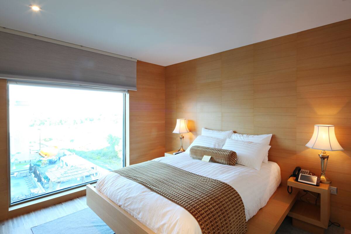 A modern hotel room with a large bed, two bedside tables with lamps, a view through a large window, and a wooden headboard.