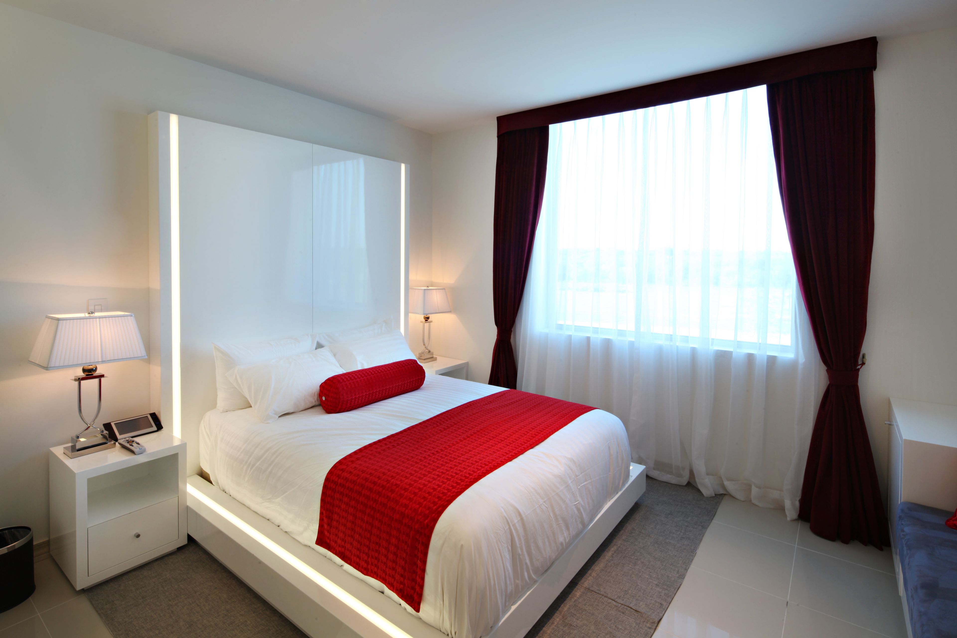A modern hotel room with a large bed, white bedding, a red throw pillow, a bedside table with a lamp, and a window with sheer curtains.
