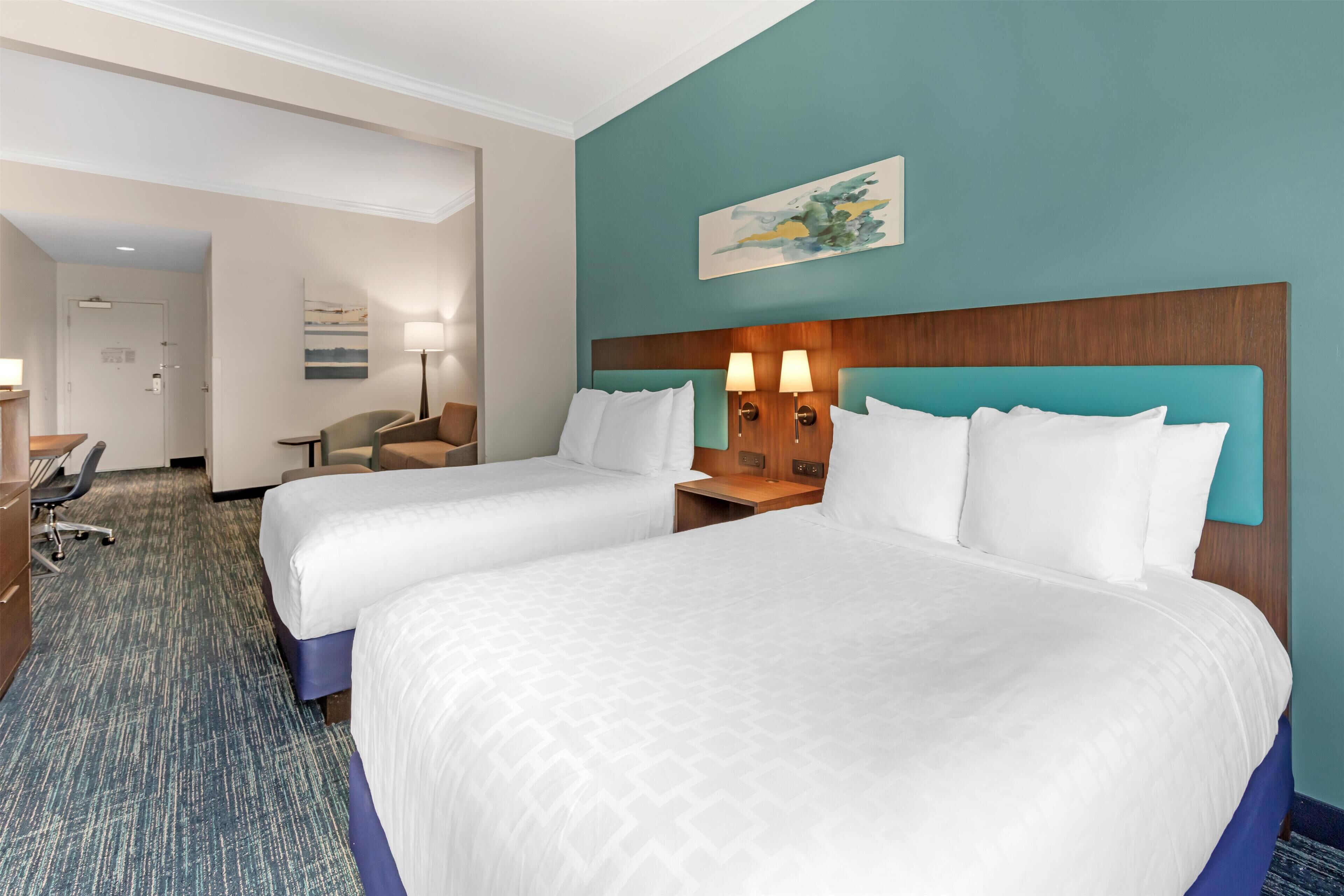 A hotel room with two beds, a desk, a chair, and a lamp.