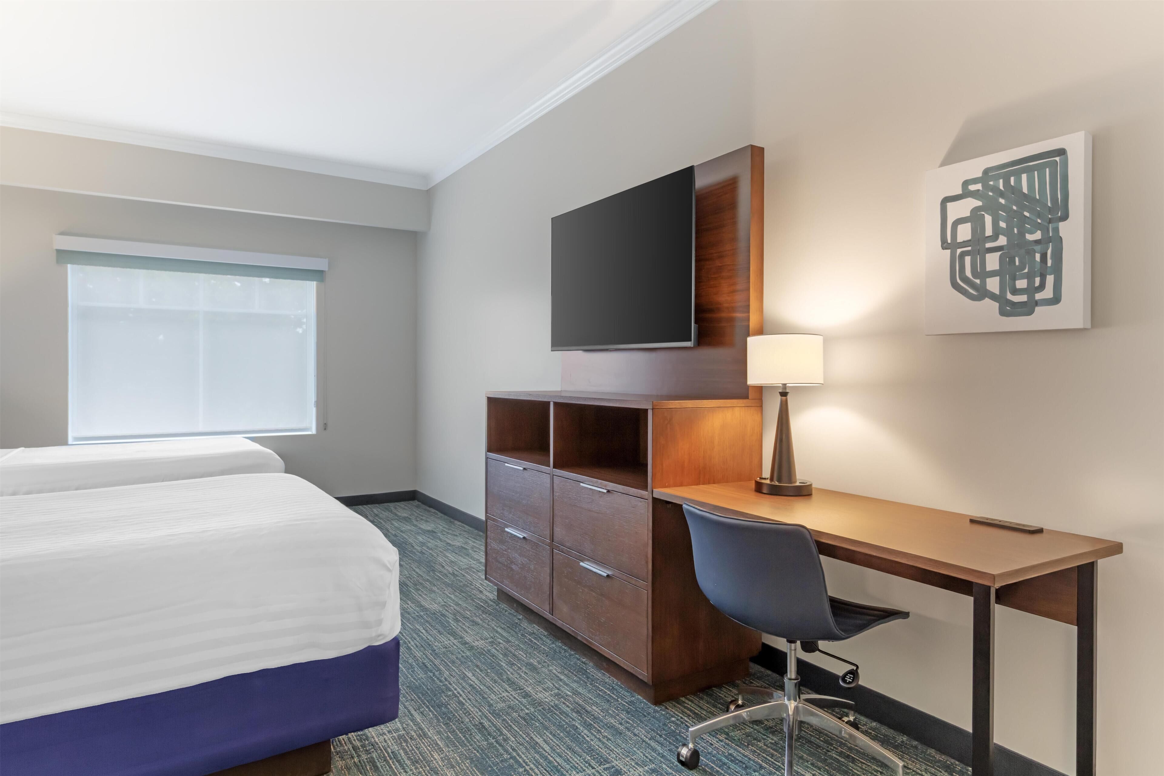 A hotel room with a bed, desk, chair, TV, lamp, and artwork on the wall.