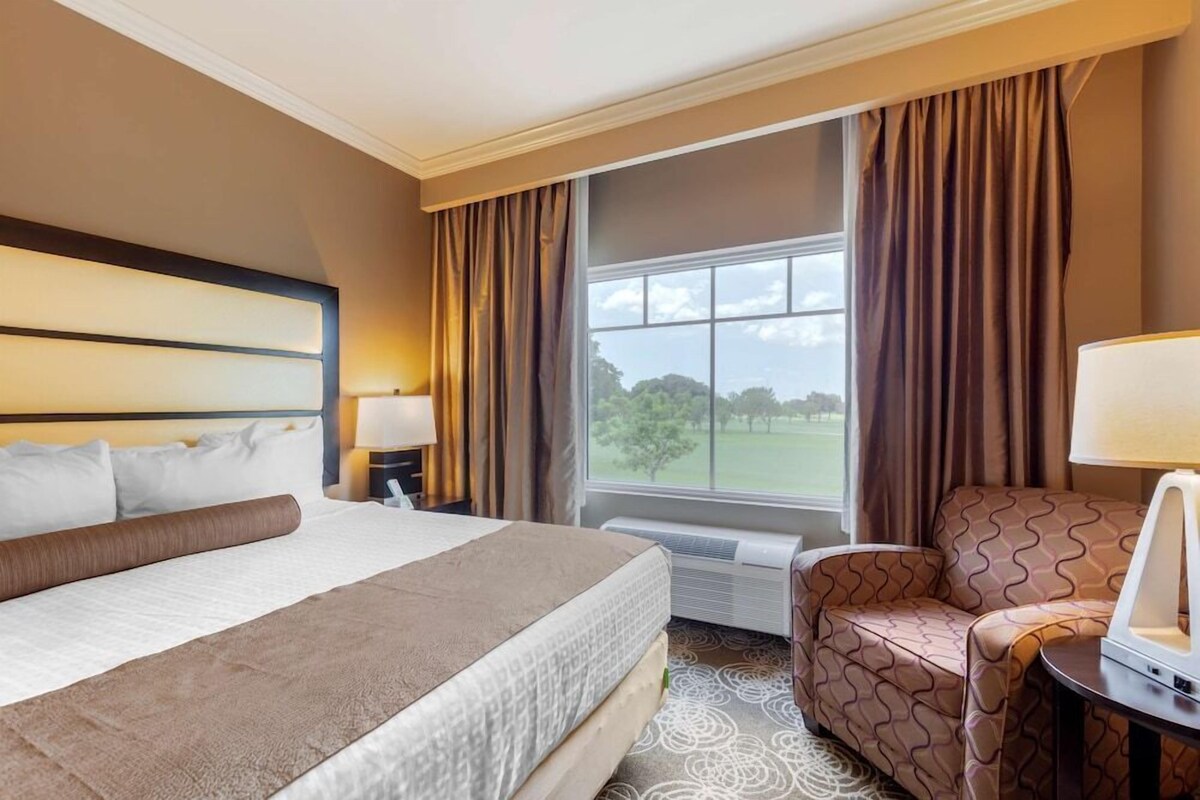 A hotel room with a large bed, a chair, a desk with a lamp, and a window with curtains.