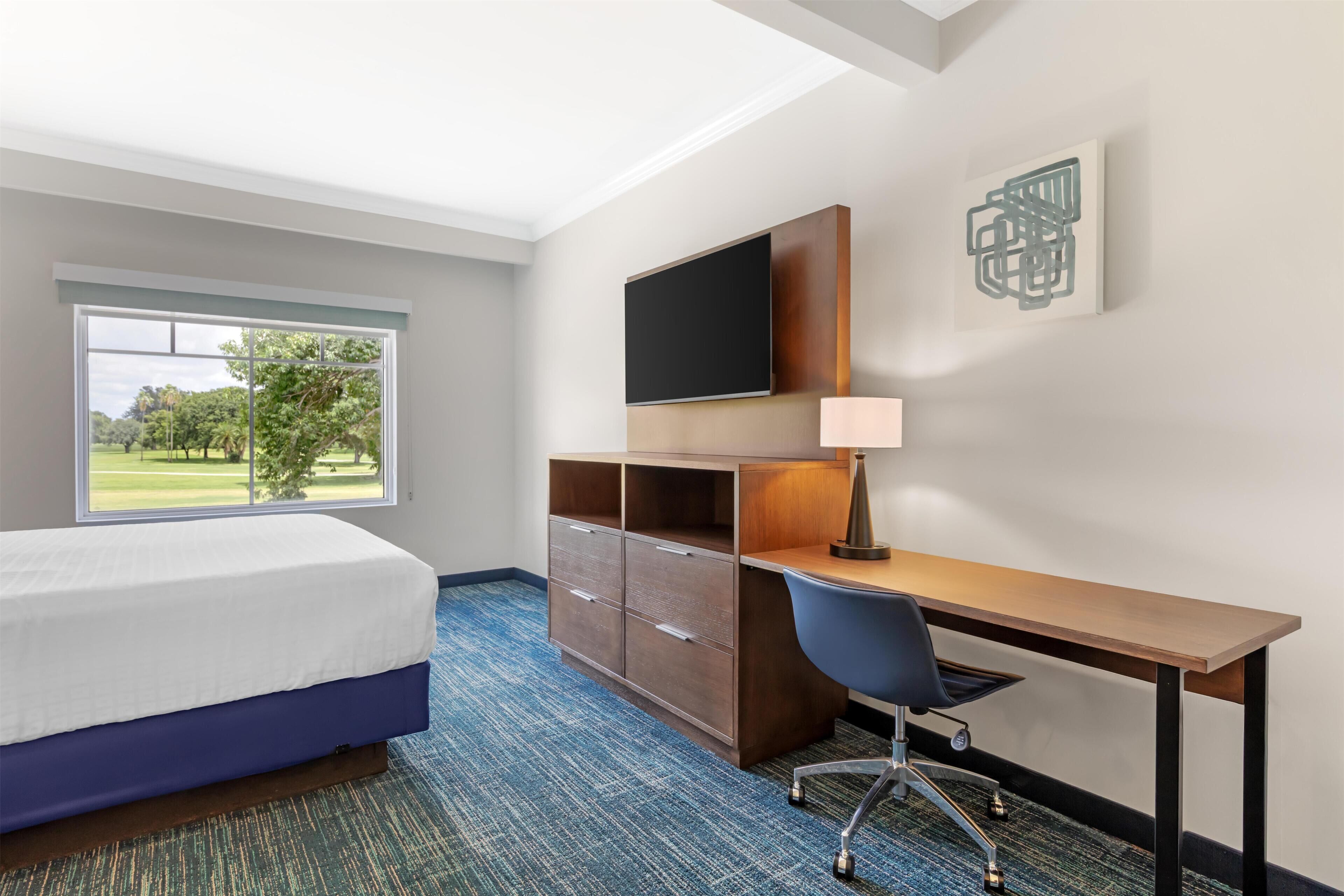 A hotel room with a bed, desk, chair, TV, and a view of a golf course.