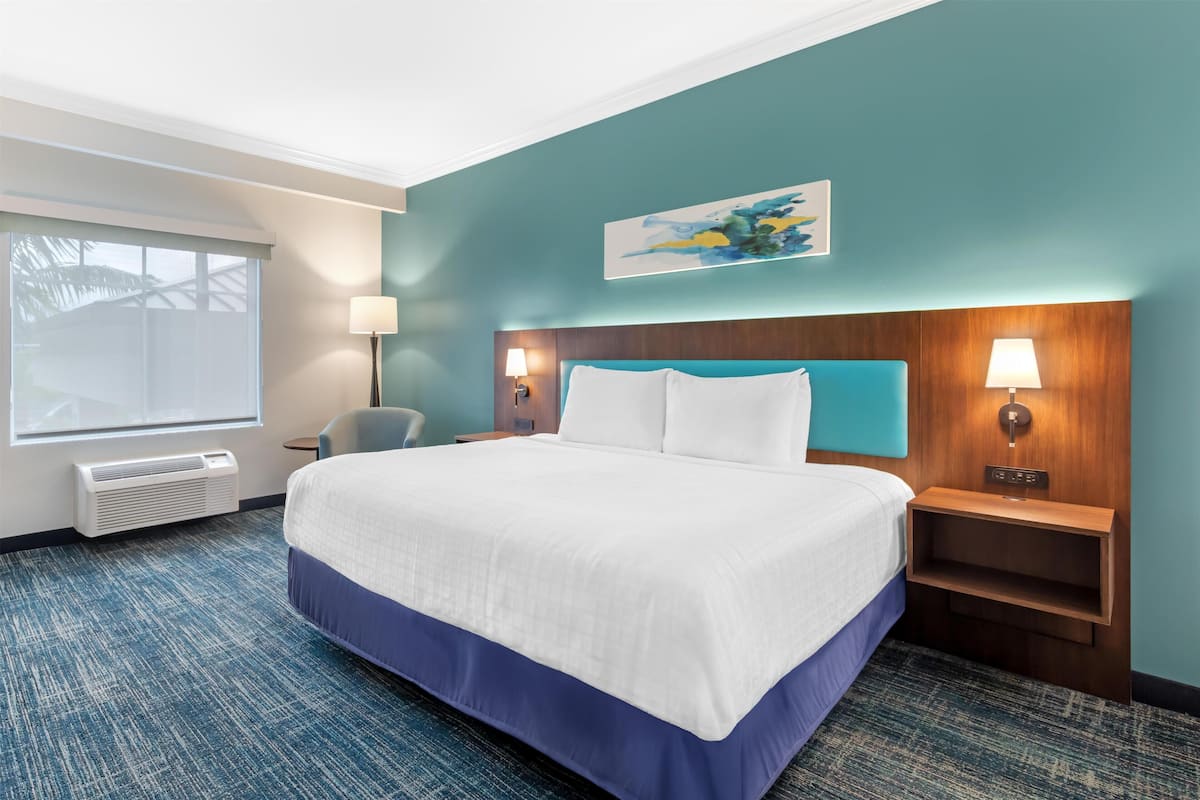 A hotel room with a large bed, two bedside tables with lamps, a window with blinds, a wall-mounted artwork, and a carpeted floor.