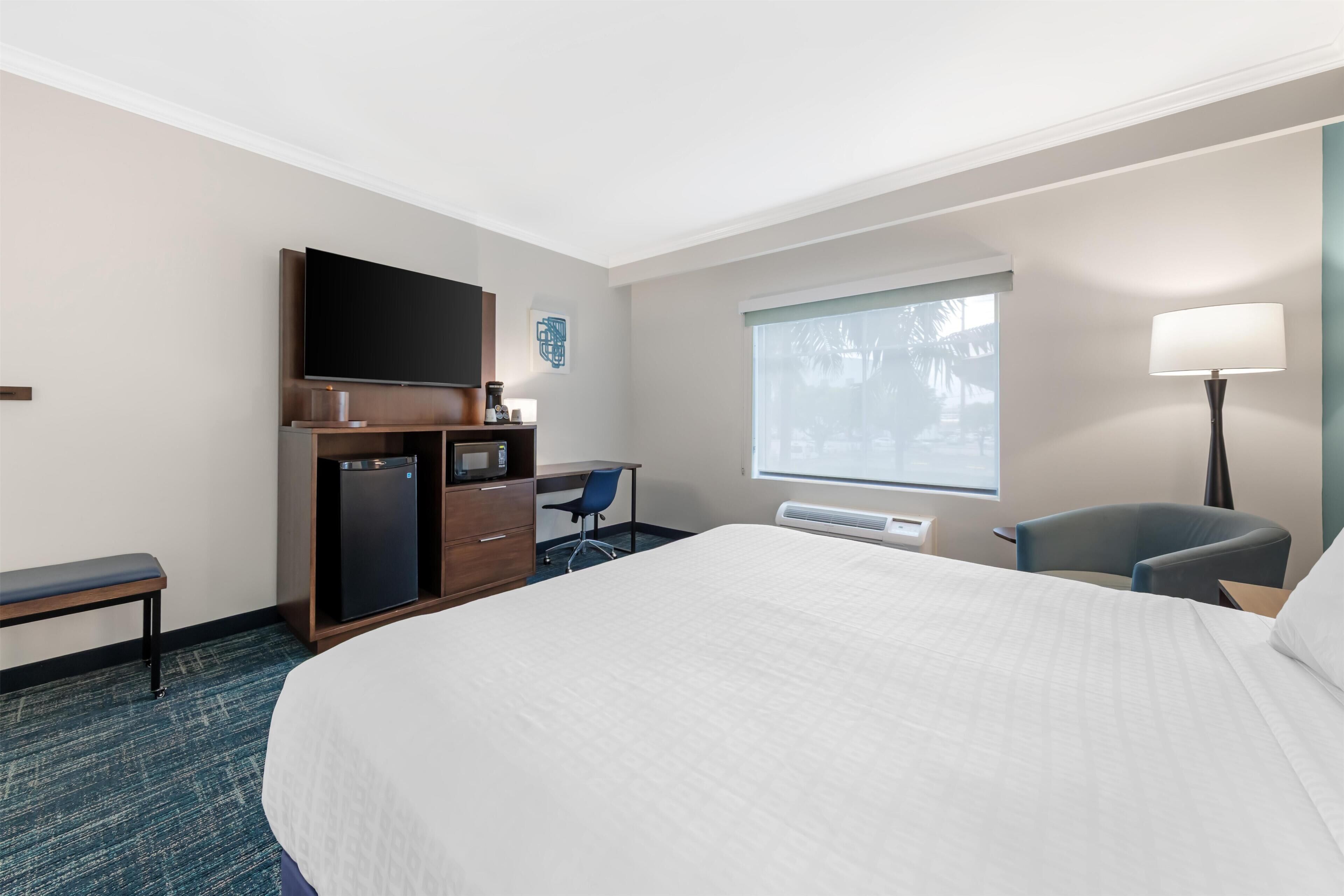 Una habitación de hotel con una cama grande, un escritorio con una silla, un televisor, un refrigerador pequeño, una lámpara y una ventana con vistas.