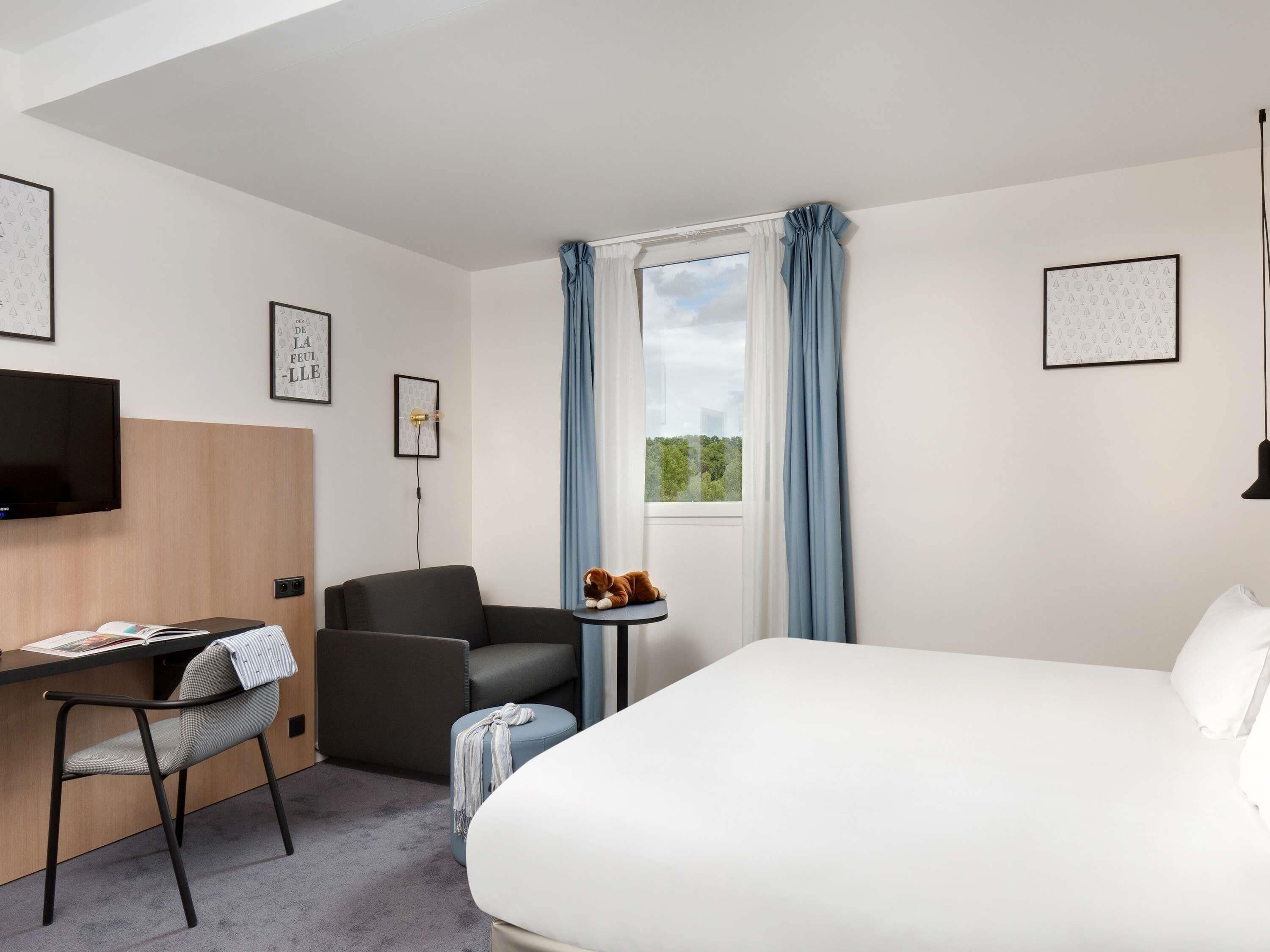 A hotel room with a bed, a desk with a chair, a television, and a small table with a cat.